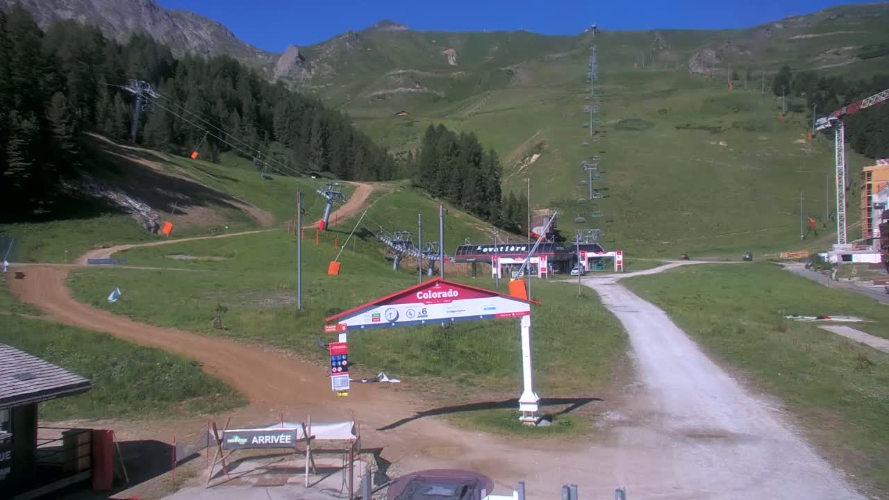 A mountainside ski resort area on a sunny day shows chairlifts, grassy hills, dirt roads, and a small building at the base of the mountain.