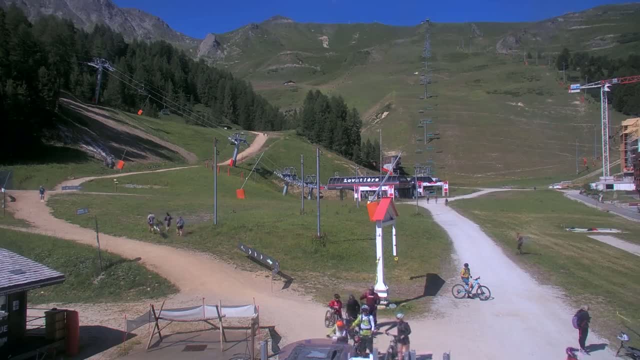 A mountainside ski resort area on a sunny day shows several people mountain biking and walking on trails near chairlifts and a small building.