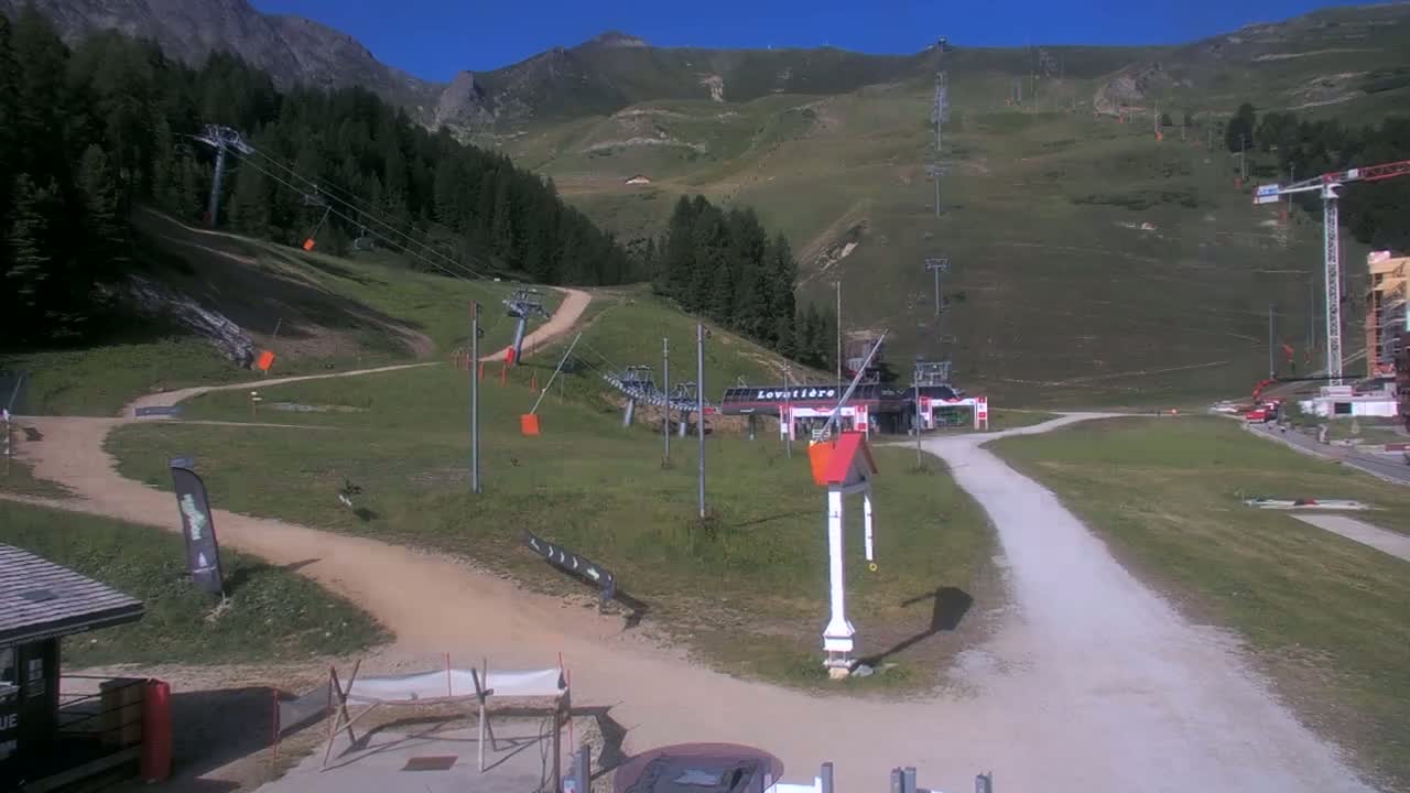 A mountainside ski resort on a sunny day shows chairlifts, grassy hills, and a dirt path leading to a building.