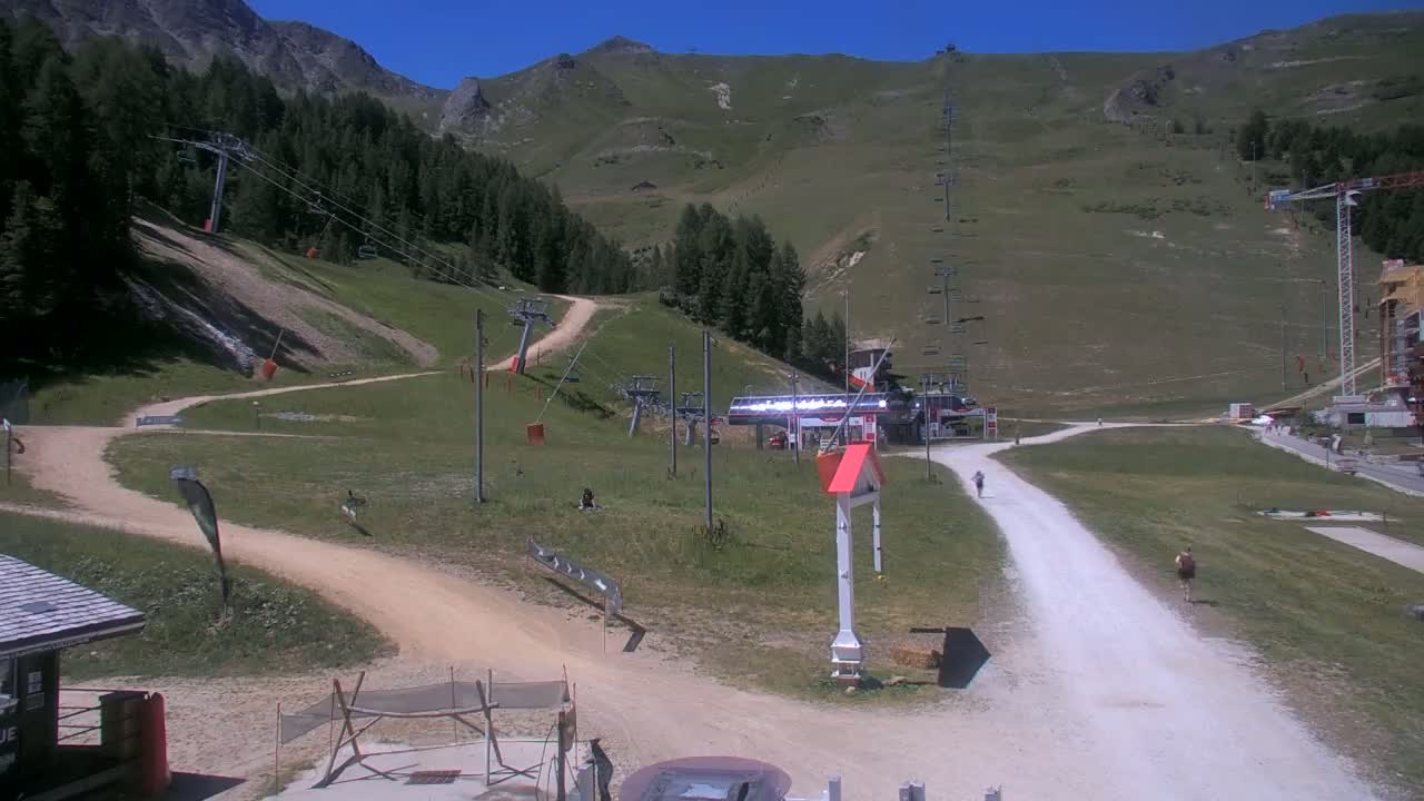 A sunny mountainside ski resort in summer shows several chairlifts, grassy hills, dirt paths, and a few people walking.