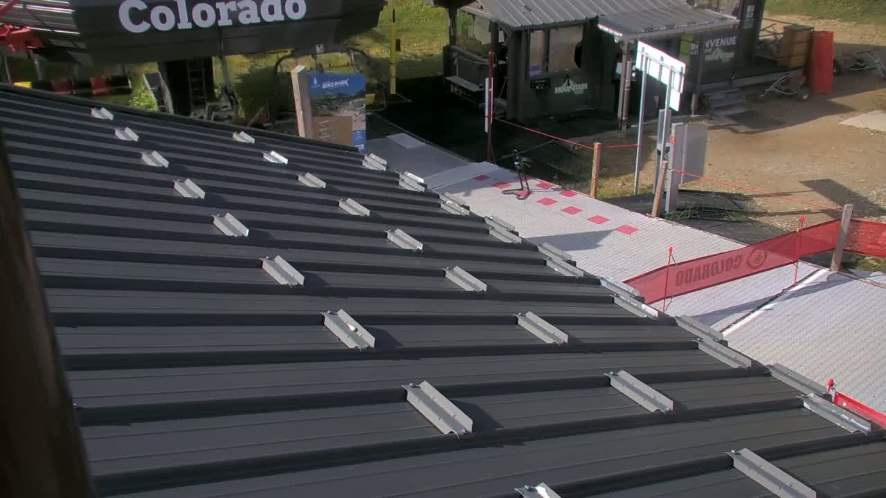 A dark gray metal roof with evenly spaced metal attachments is shown, overlooking an outdoor area with a light gray walkway and red safety barriers on a sunny day.