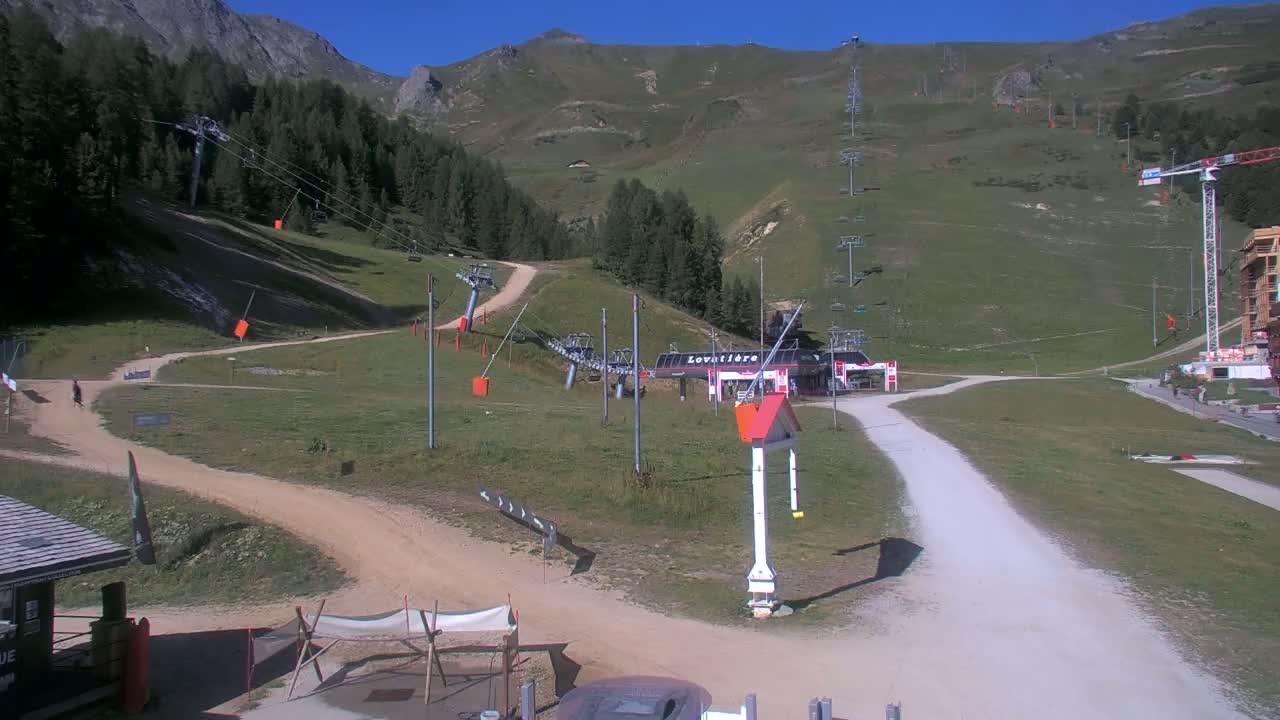 A sunny mountainside scene shows a ski lift station with several chairlifts and pathways winding through grassy hills, with a construction crane visible in the background.
