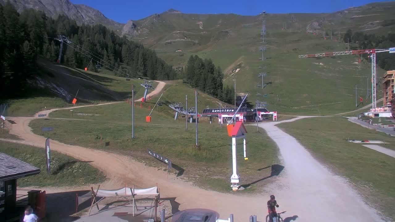 A mountainside ski resort in sunny weather shows several chairlifts, dirt paths, and a person riding a bicycle.