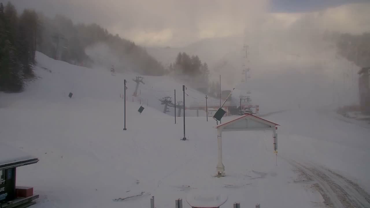 A snow-covered ski slope featuring multiple ski lifts and active snowmaking cannons is shrouded in a foggy haze under an overcast sky.
