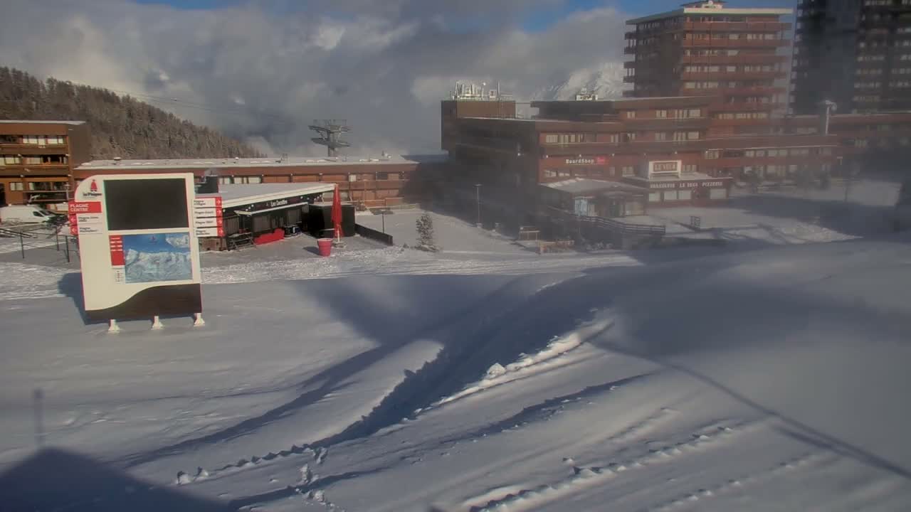A snow-covered ski resort village with multi-story brown buildings and an information board is visible under a partly cloudy sky, with atmospheric haze or light blowing snow obscuring parts of the right side.