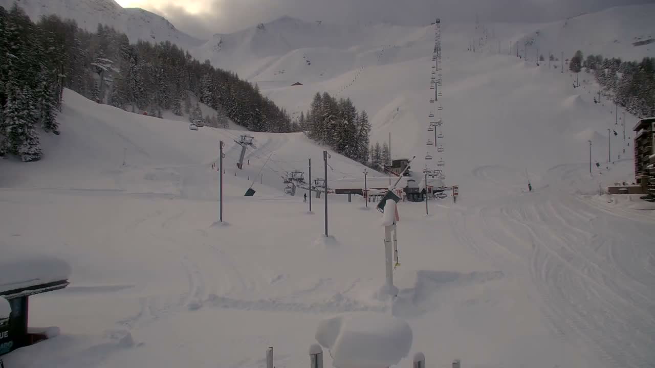 A snow-covered ski resort features wide slopes with ski tracks, scattered evergreen trees, several ski lifts including a prominent tall one, and some buildings nestled against the base of distant, hazy mountains, all under a bright, overcast sky.