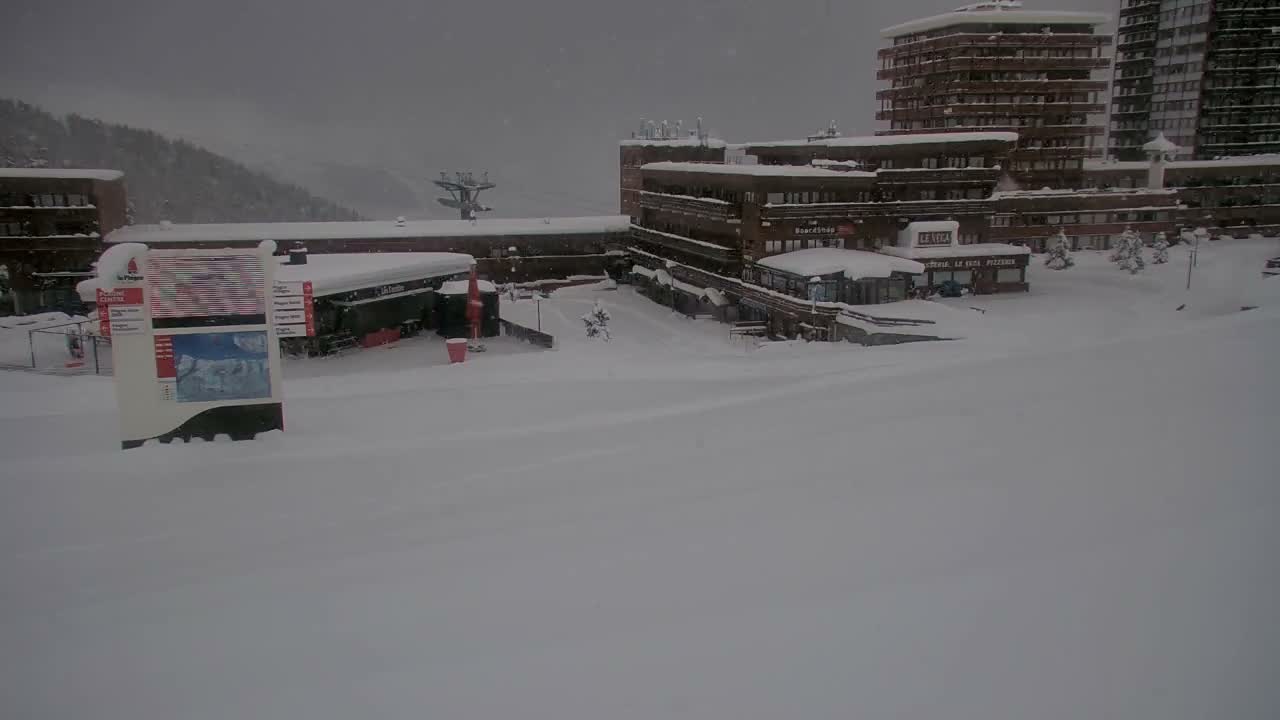 A snow-covered multi-building complex, likely a ski resort, is blanketed under a heavy layer of fresh snow with an overcast, grey sky and visible snowfall.