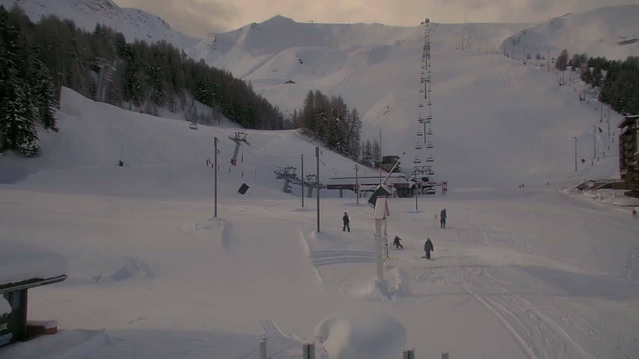 A wide shot of a snow-covered ski resort under cloudy skies, featuring multiple ski lifts, sparse pine trees, distant mountains, and a few skiers on the slopes.