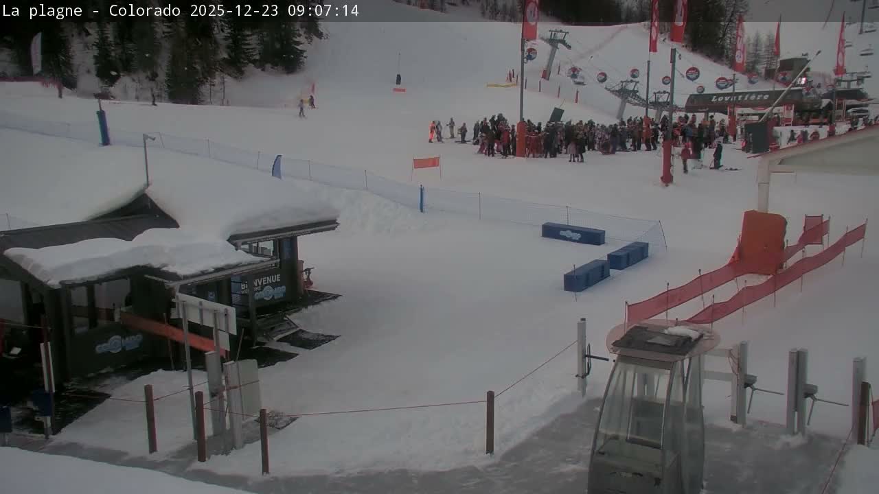 The image depicts a bustling ski resort covered in snow on an overcast day, with numerous skiers and snowboarders gathered near ski lifts and buildings on the slopes.