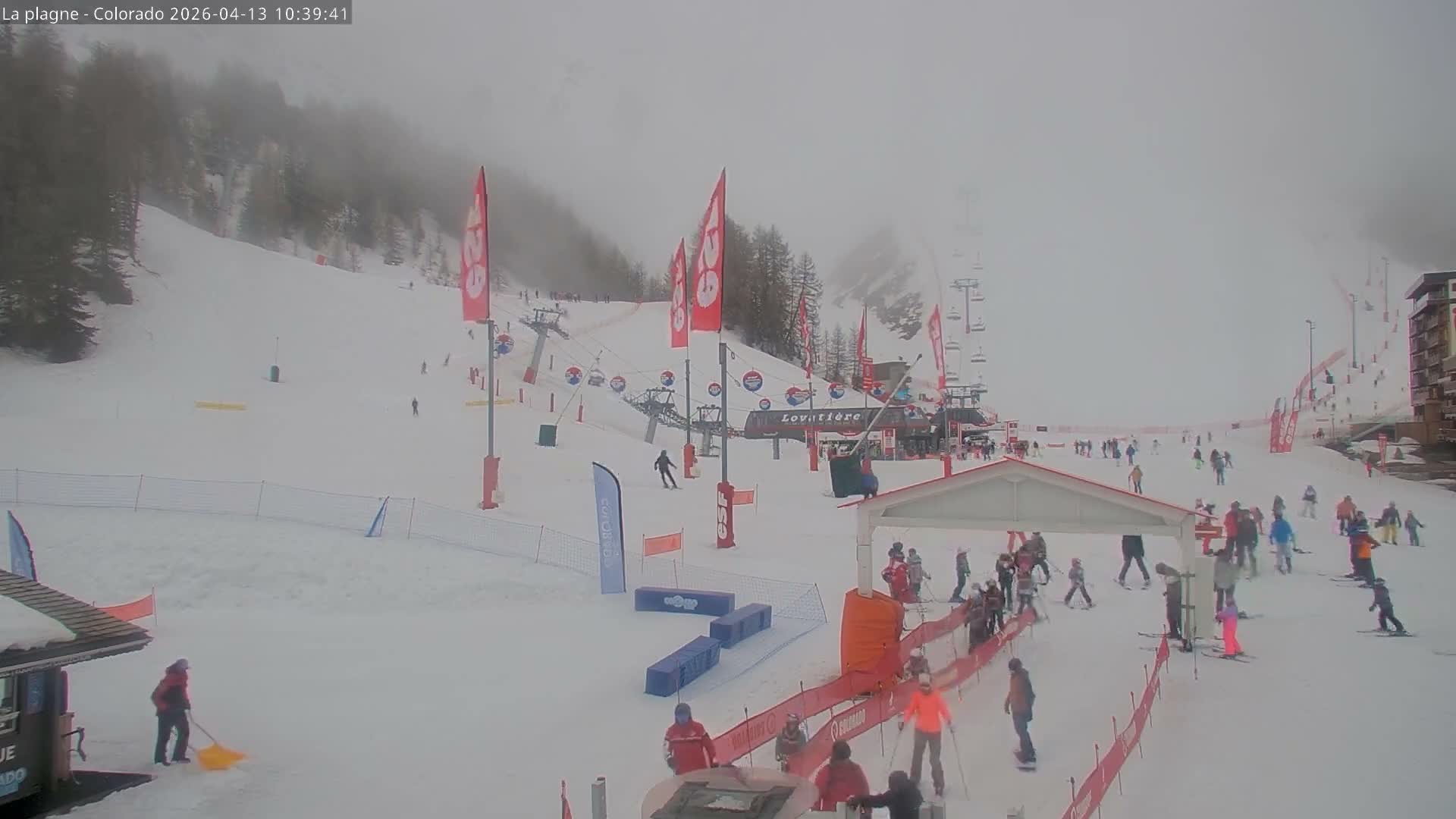 The image depicts a bustling ski resort covered in snow on an overcast day, with numerous skiers and snowboarders gathered near ski lifts and buildings on the slopes.