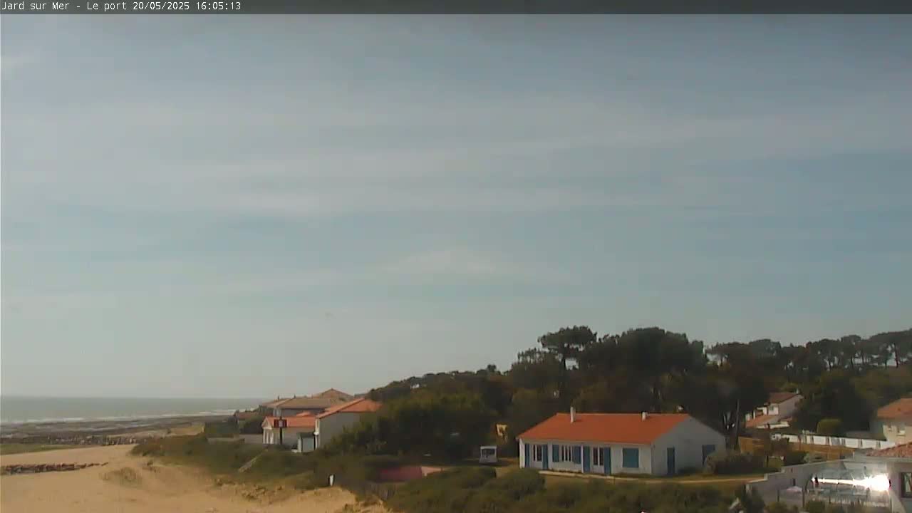 A mostly sunny day shows a calm ocean, sandy beach, and several houses nestled among green trees near the coast.