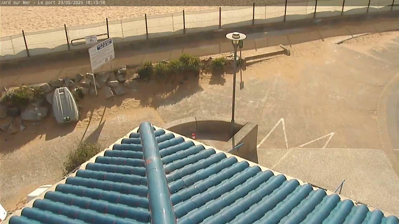A blue tiled roof overlooks a sandy area with a small boat and a lamppost, near a fence and beach.