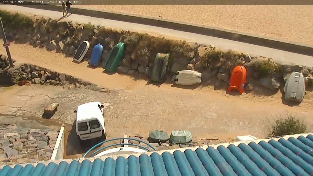 A white van is parked near a rocky shore where several small boats are beached in muddy water under a sunny sky.