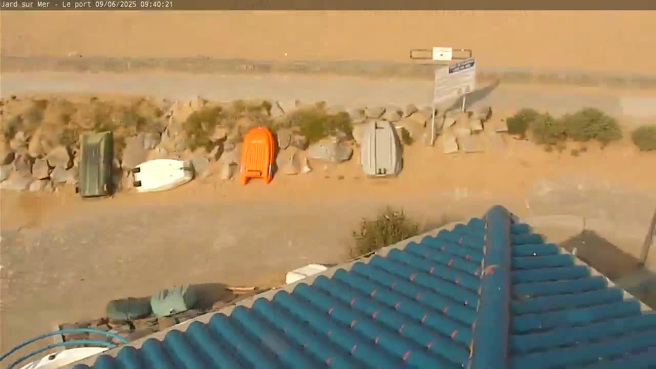 A view from a rooftop shows several small boats and containers resting on a sandy, rocky area next to a paved path on a sunny day.