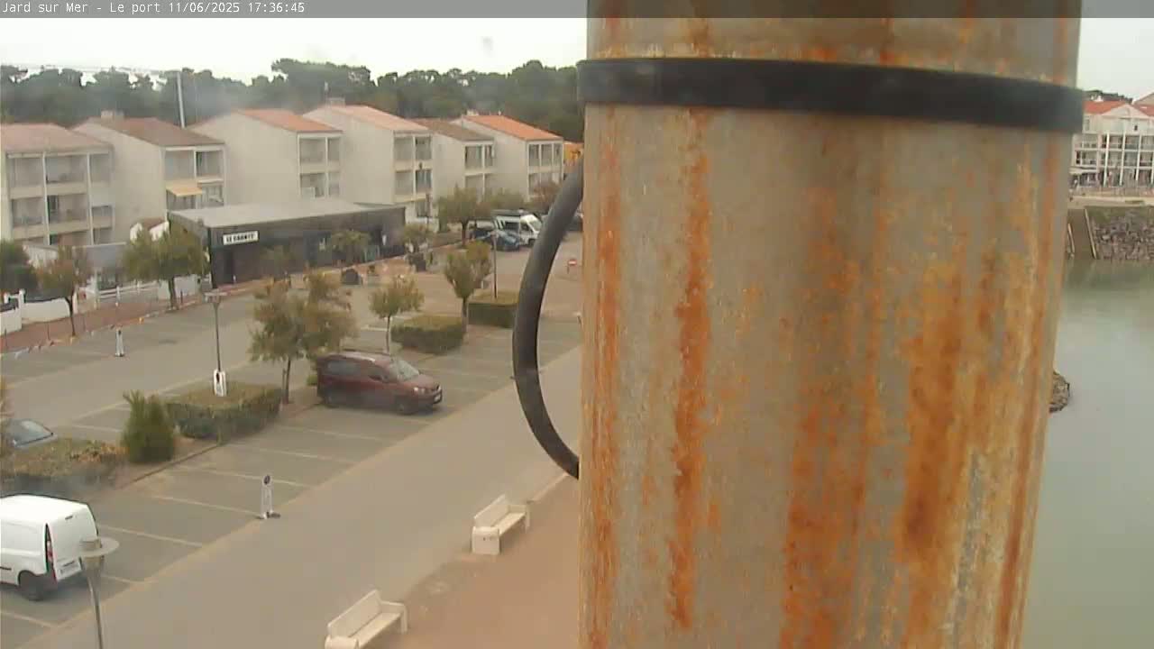 An overcast day shows a parking lot next to apartment buildings and a body of water, all viewed from a high vantage point near a rusty metal pole.