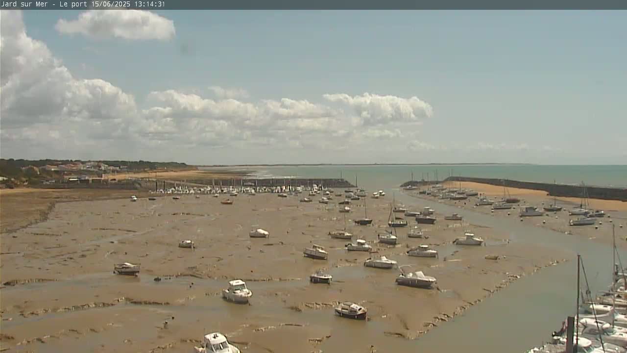 Under a partly cloudy sky, numerous boats are grounded in a shallow, muddy harbor.