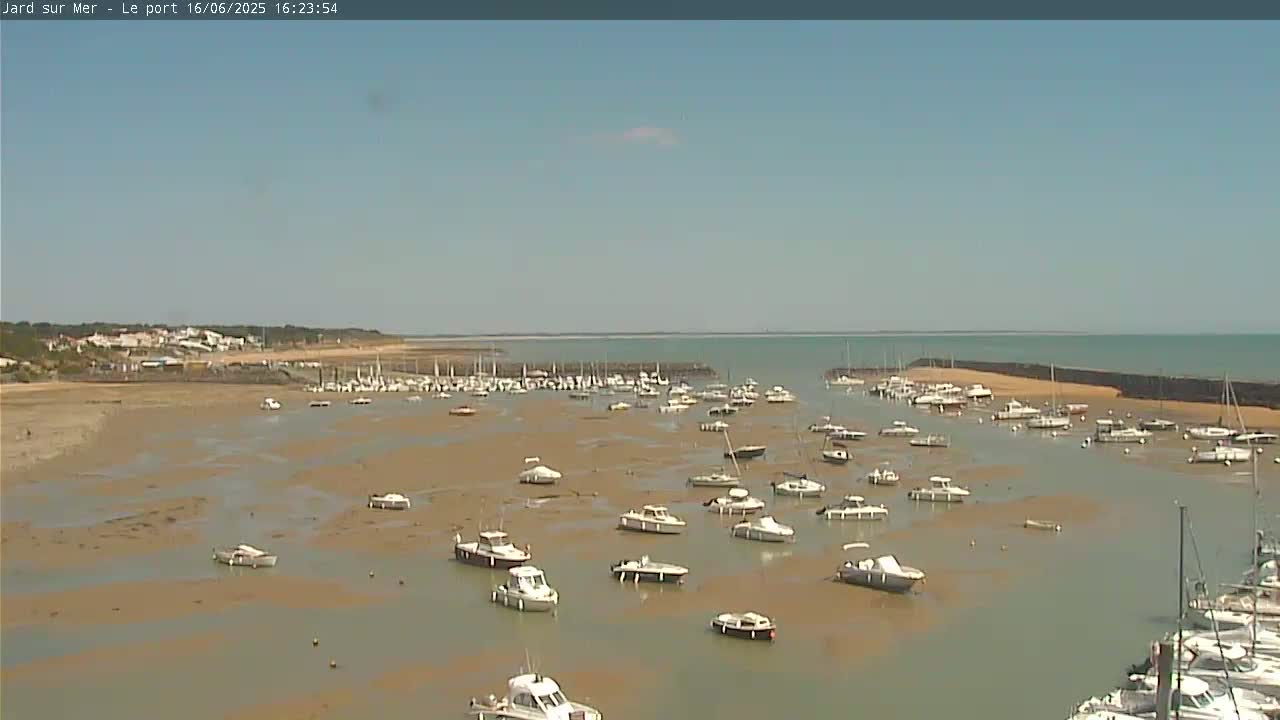 A mostly sunny day shows numerous boats in a harbor at low tide, with a calm sea and sandy shoreline visible in the distance.