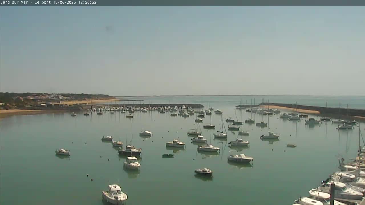 A calm, sunny day reveals a harbor filled with numerous boats and yachts, nestled beside a sandy shoreline and a small town in the distance.