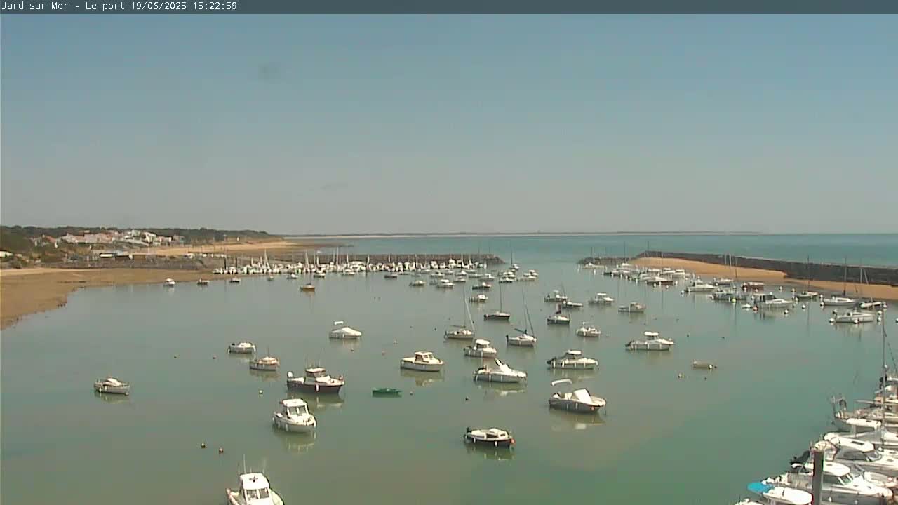 A calm, sunny day shows a harbor filled with numerous small boats,  with a sandy beach and distant coastline visible.