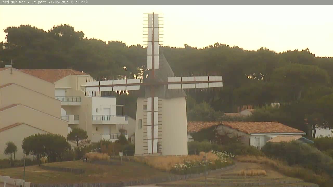 A windmill stands in a grassy area near tan buildings and a line of dark green trees under a hazy sky.