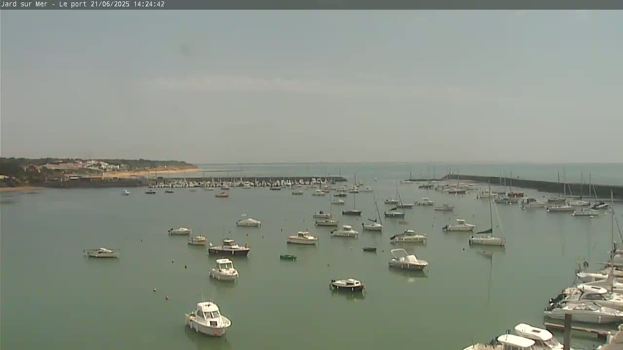 A calm, light-grey day reveals a harbor filled with numerous sailboats and motorboats, nestled against a shoreline with buildings and sandy beaches.