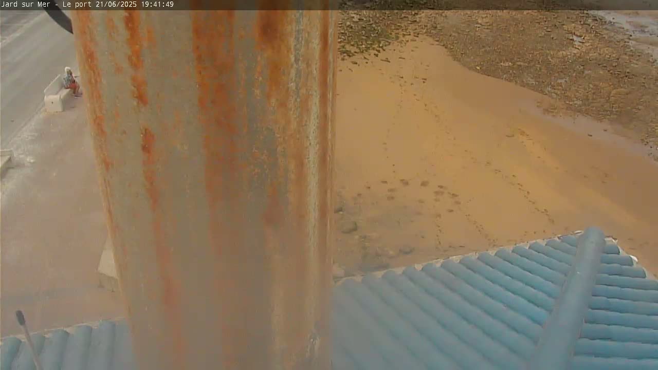 A rusty metal pole is seen next to a teal roof overlooking a sandy beach with footprints.