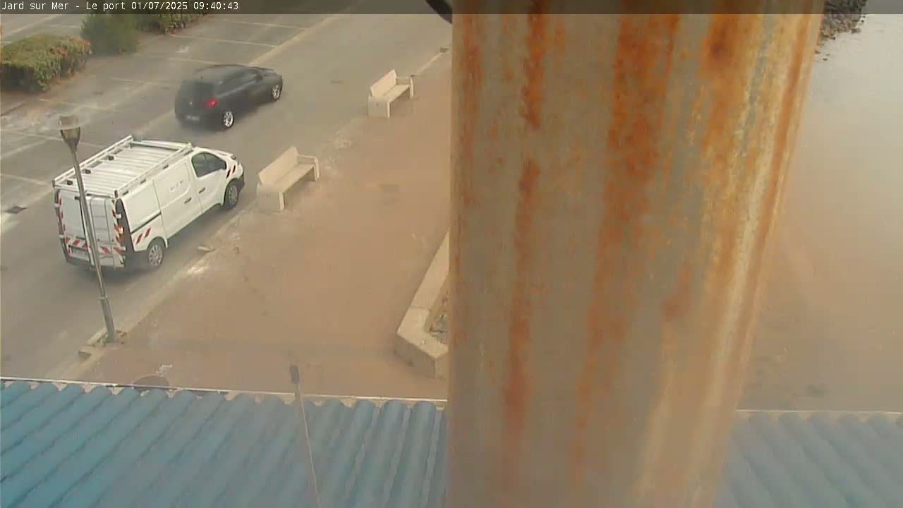 A flooded parking lot with two vehicles and park benches is viewed from above a rusty pole.