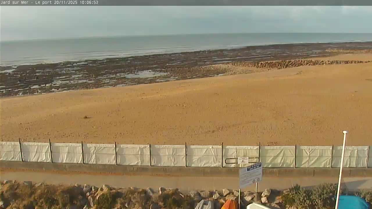 A wide sandy beach, backed by a rocky intertidal zone and the ocean under an overcast sky, is separated by a white barrier from a foreground of rocks and sparse vegetation.