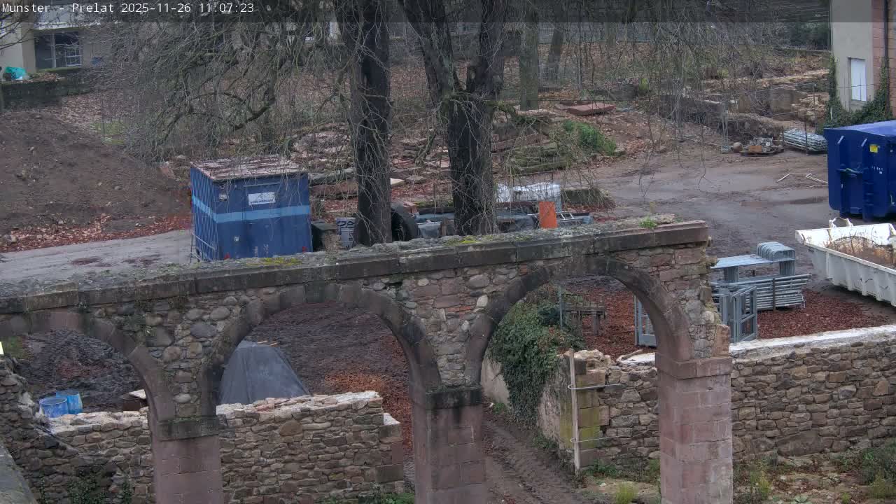 The image displays a muddy, debris-strewn construction site on an overcast day, featuring a stone archway in the foreground, several bare trees, and various blue containers and construction materials scattered across the midground.