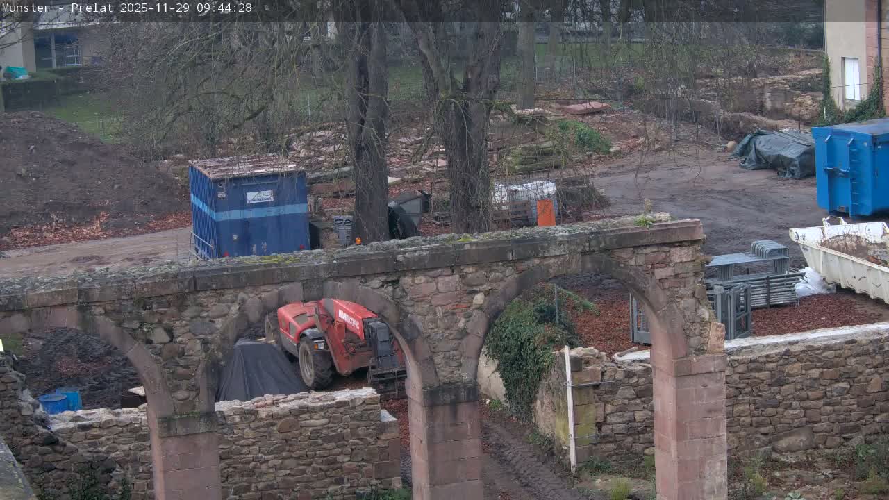 A construction site is depicted under an overcast sky, featuring a prominent stone archway, a red telehandler, and various building materials scattered across muddy ground with bare trees in the background.
