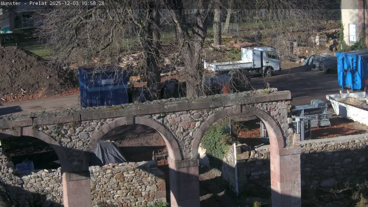 Under clear, sunny skies, a stone arch bridge stands in the foreground, overlooking a dirt area with bare trees, a white truck, and various blue containers, indicating a construction or storage site.