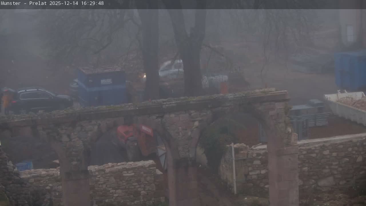 Under clear, sunny skies, a stone arch bridge stands in the foreground, overlooking a dirt area with bare trees, a white truck, and various blue containers, indicating a construction or storage site.
