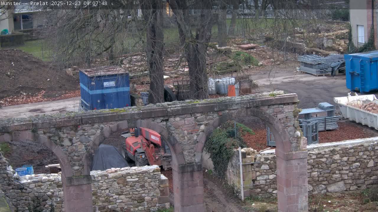 An outdoor construction or renovation site is depicted on an overcast day, featuring prominent stone arches in the foreground, an orange telehandler, blue containers, and stacks of metal scaffolding amidst bare trees and disturbed earth.