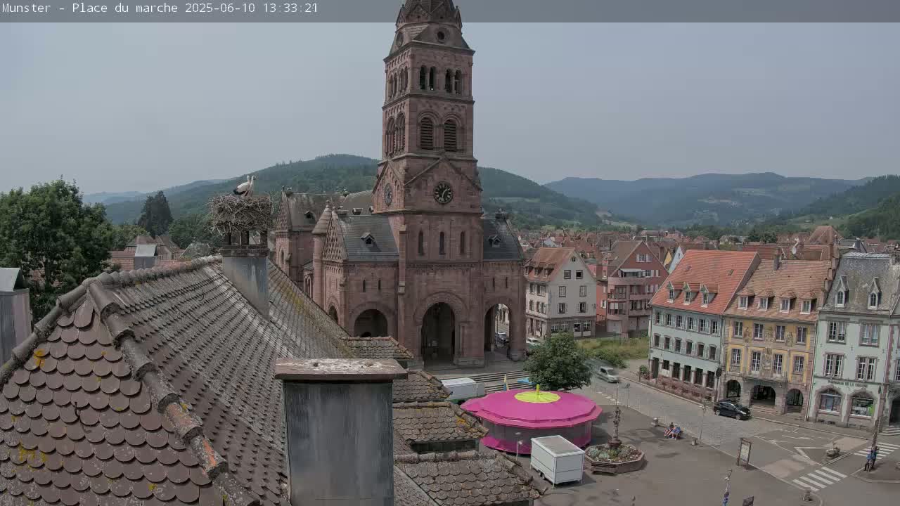 A town square with a large church and several buildings, a carousel, and a stork's nest on a chimney, all under a mostly cloudy sky.