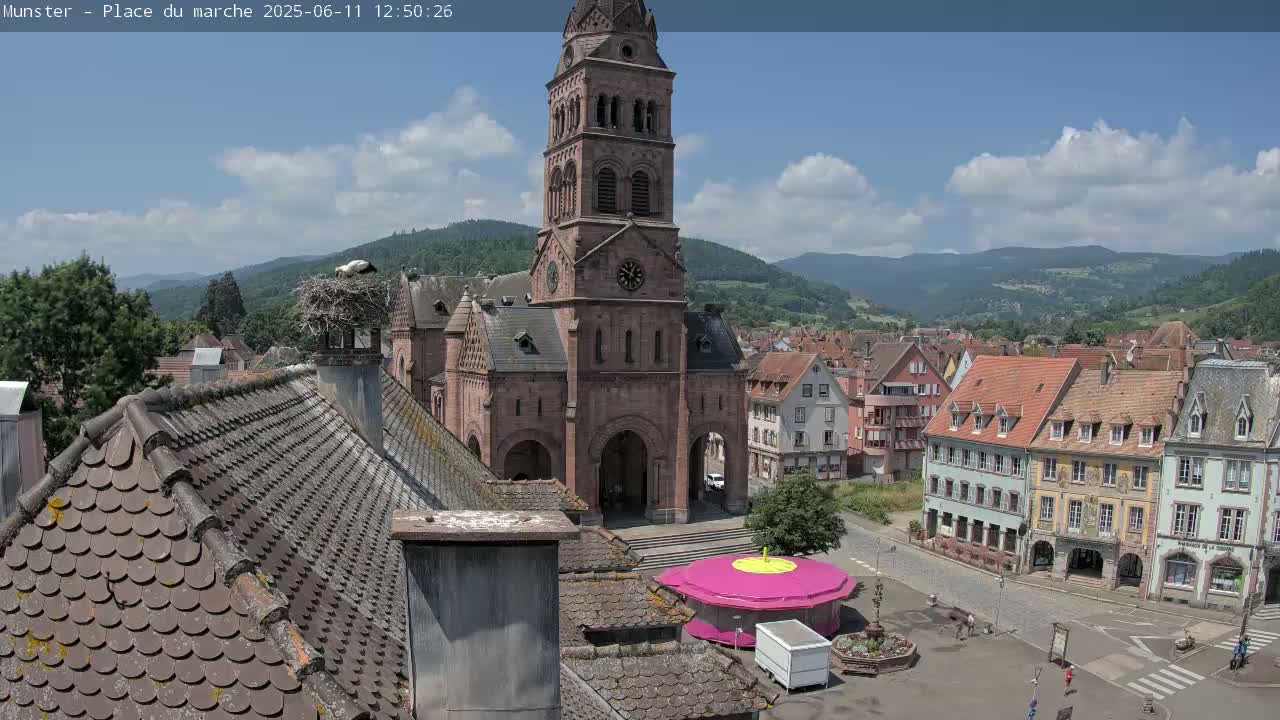 A partly cloudy day reveals a town square with a church and surrounding buildings, a stork's nest atop a chimney, and a small carousel in the foreground, nestled in a valley surrounded by mountains.