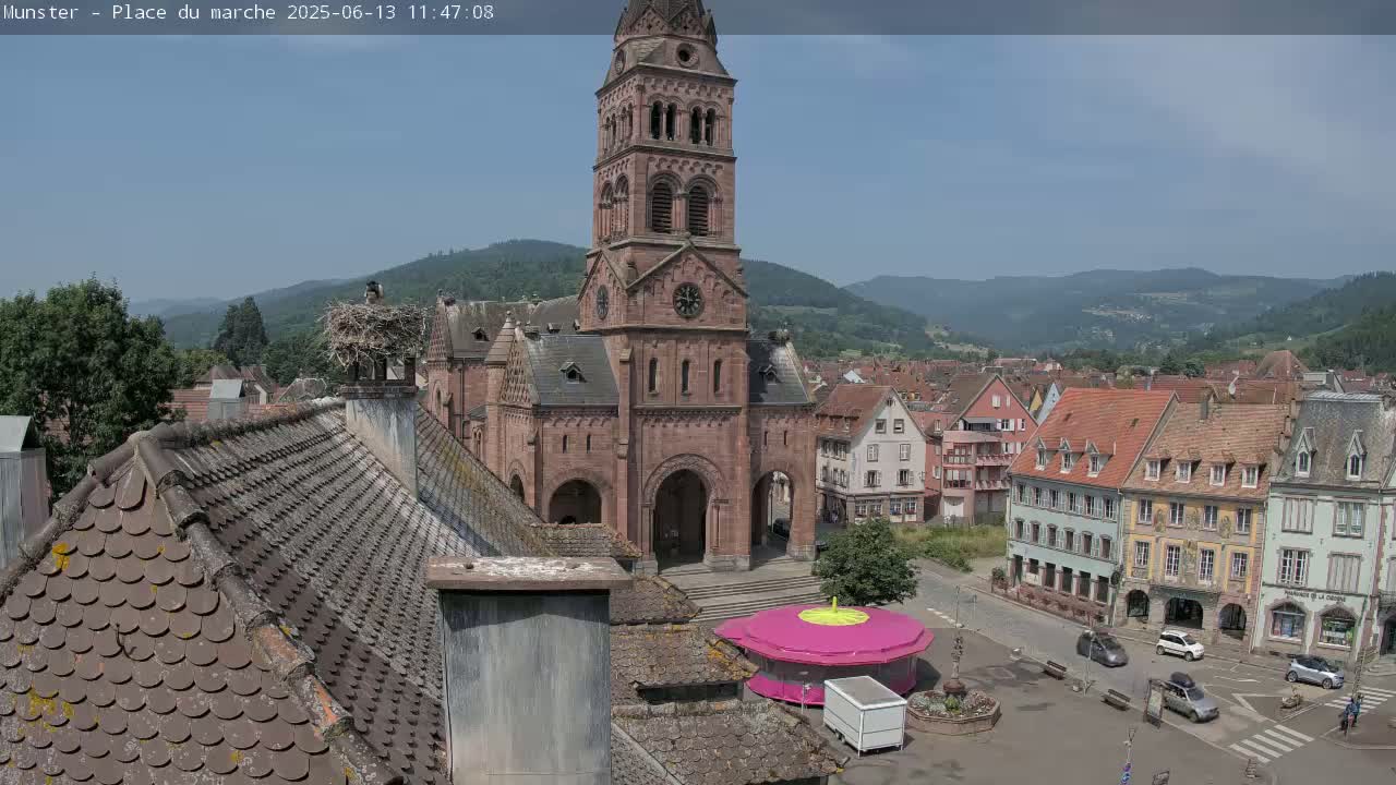 A large church with a tall tower stands in a town square on a sunny day, surrounded by other buildings, with mountains visible in the background and a stork's nest on a chimney.