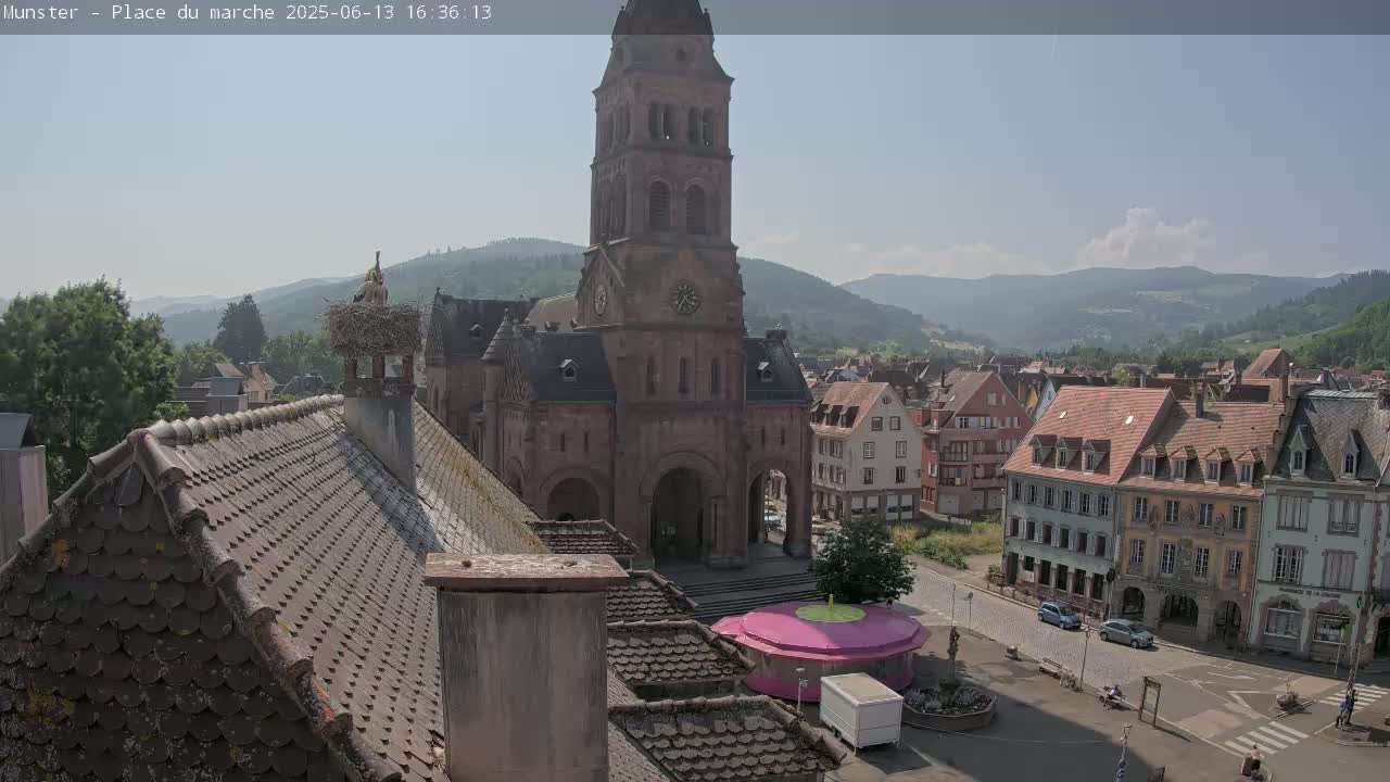 A high-angle, sunny view shows a town square with a large church, surrounding buildings, and a stork's nest on a chimney, all set against a backdrop of rolling hills.