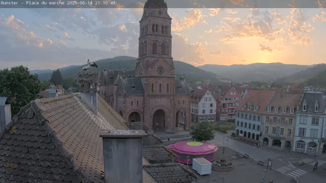 A partly cloudy day reveals a town square dominated by a large stone church, with storks nesting on a nearby chimney, and surrounded by traditional buildings nestled against a backdrop of rolling hills.