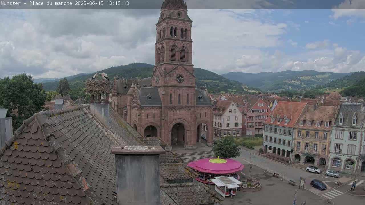 A partly cloudy day reveals a town square with a large church, several buildings, and a small carousel, all nestled in a valley surrounded by mountains; a stork's nest sits atop a chimney.