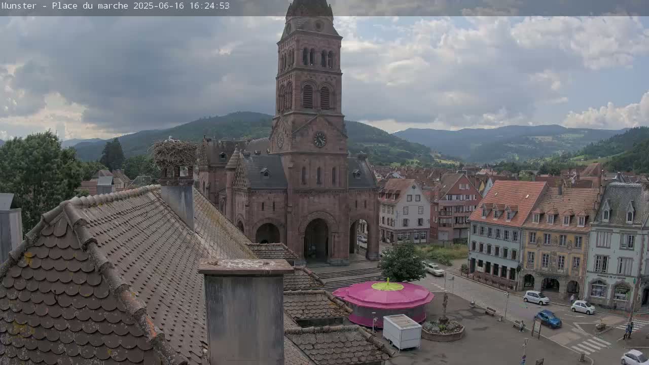 A partly cloudy day shows a town square with a large church, surrounding buildings, a small carousel, and a stork's nest on a chimney, all nestled in a valley between mountains.