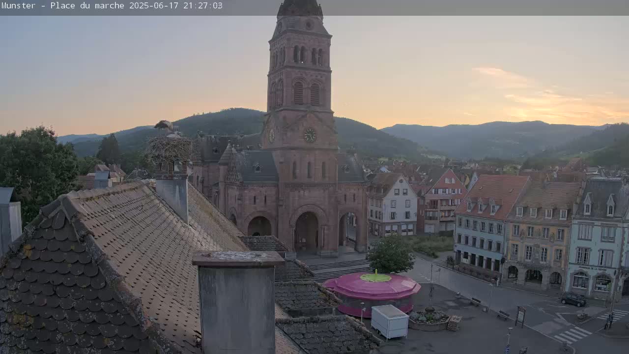 A view of a town square at sunset, featuring a tall church, several buildings, a stork's nest on a chimney, and a carousel.
