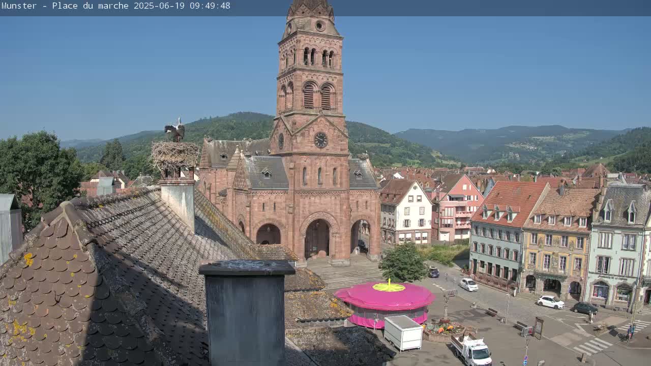 A church with a tall tower is situated in a town square overlooked by hills under a clear blue sky, with storks perched on a nest atop a chimney.