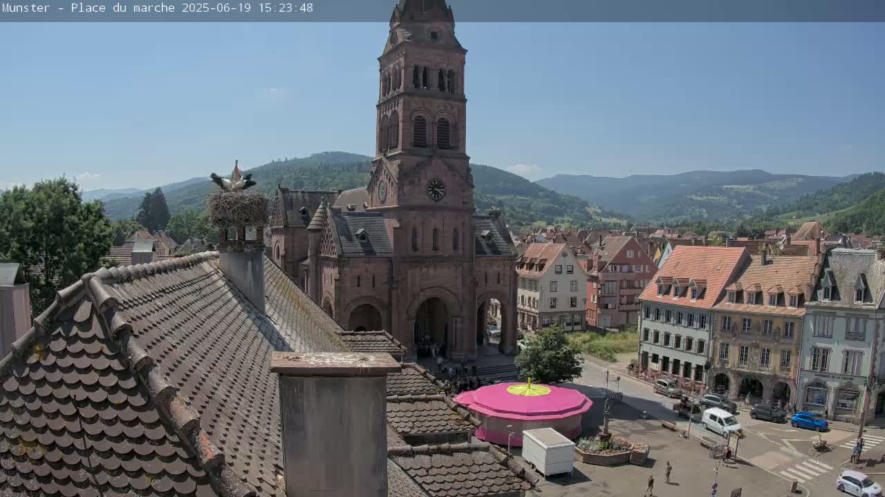 A high-angle view of a town square on a sunny day, showcasing a church with a tall tower, surrounding buildings, and a small pink carousel, with storks perched on a nearby rooftop.