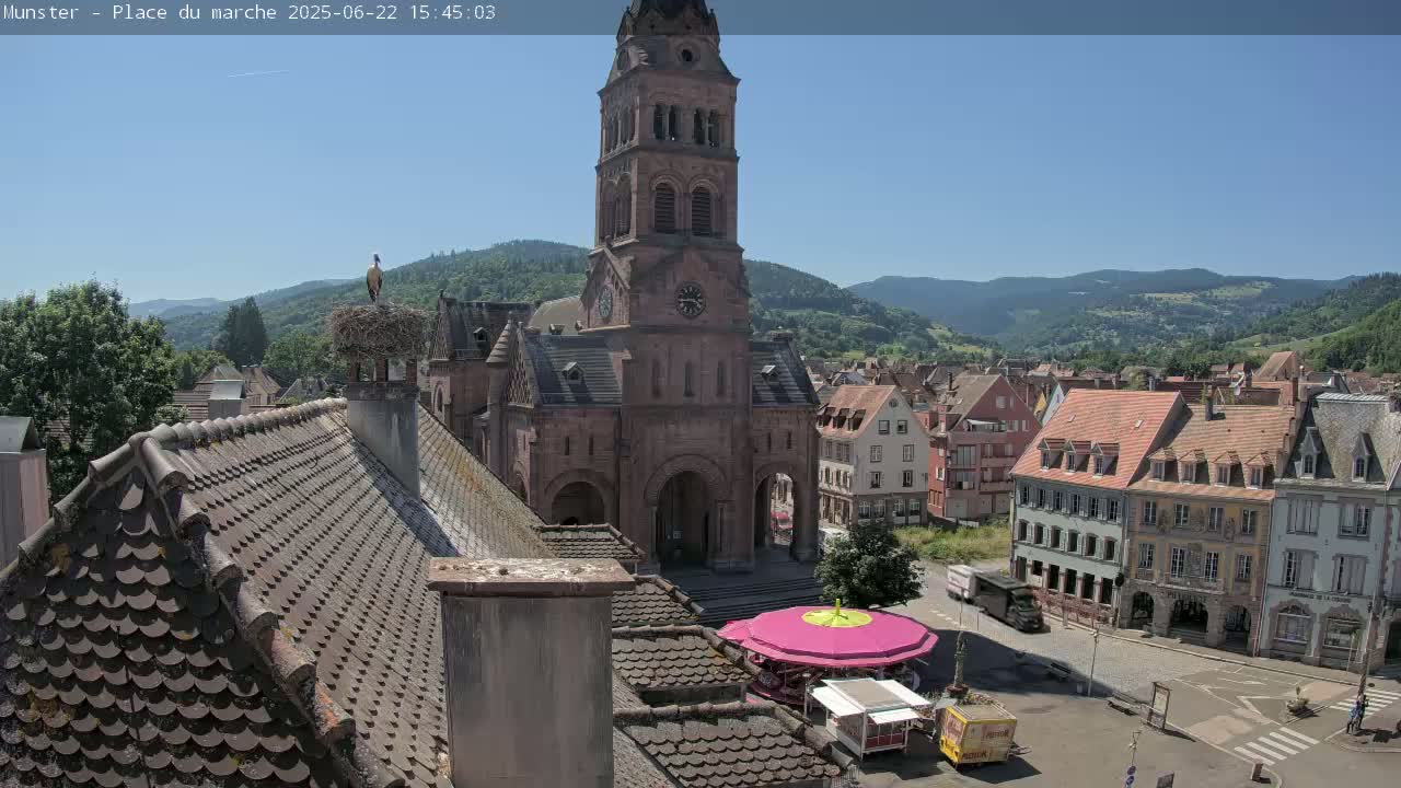 A high-angle, sunny view of a town square dominated by a large church with a tall tower, a small carousel, and surrounding buildings, all nestled in a valley under a clear blue sky.