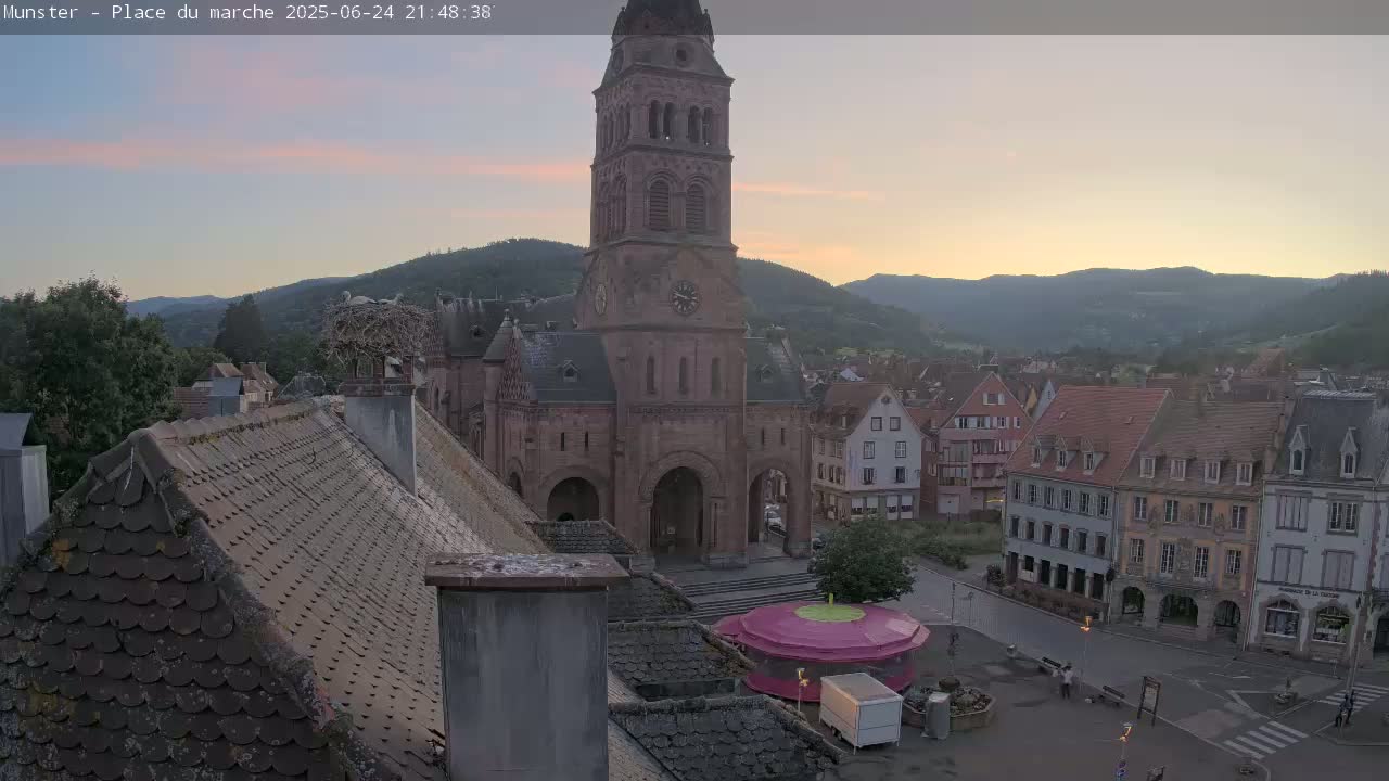 A view of a town square at sunset, featuring a large church, surrounding buildings, a stork's nest on a chimney, and a small carousel in the square.
