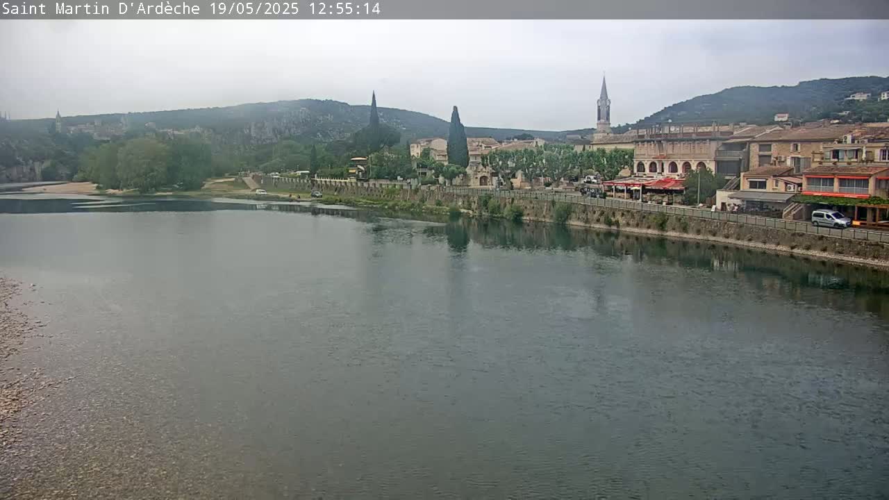 A river flows past a village nestled in a valley under an overcast sky.