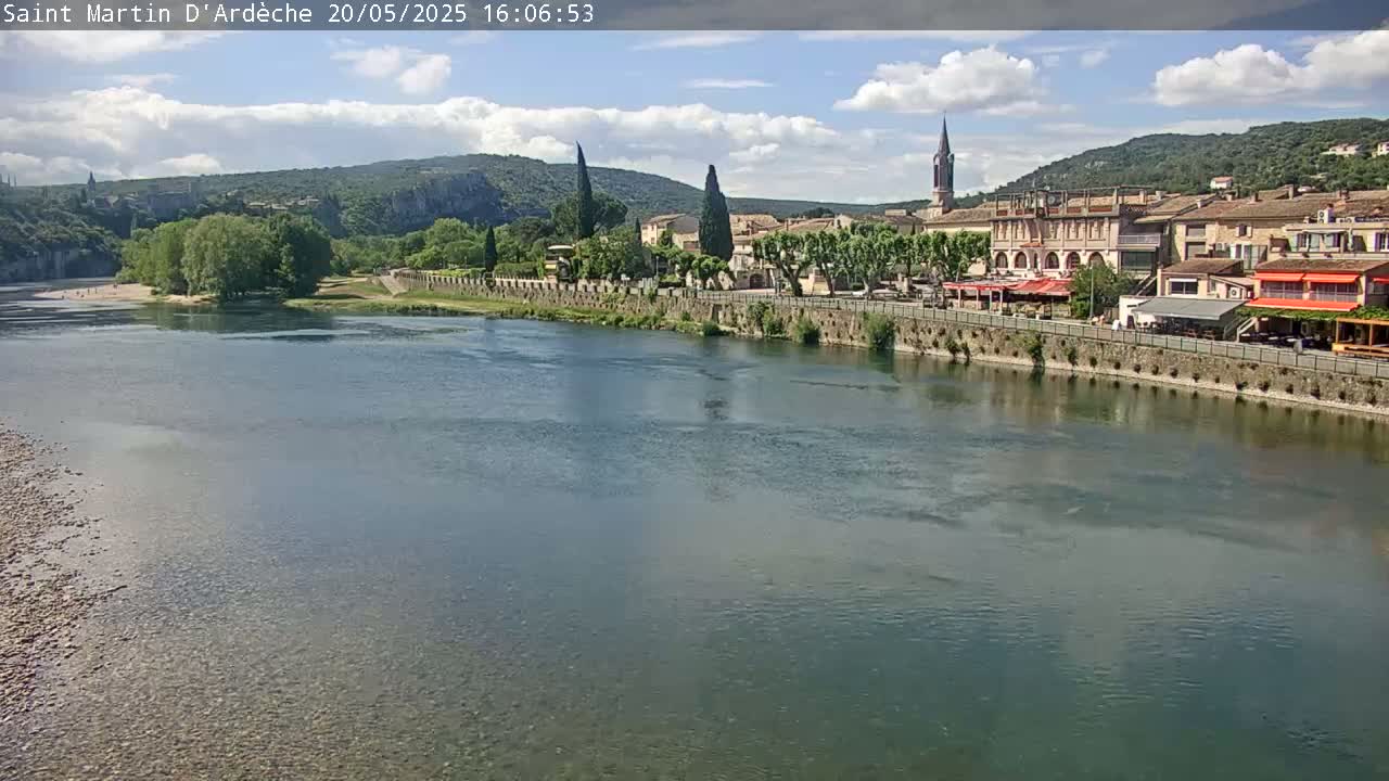 A river flows past a village nestled against hills under a partly cloudy sky.