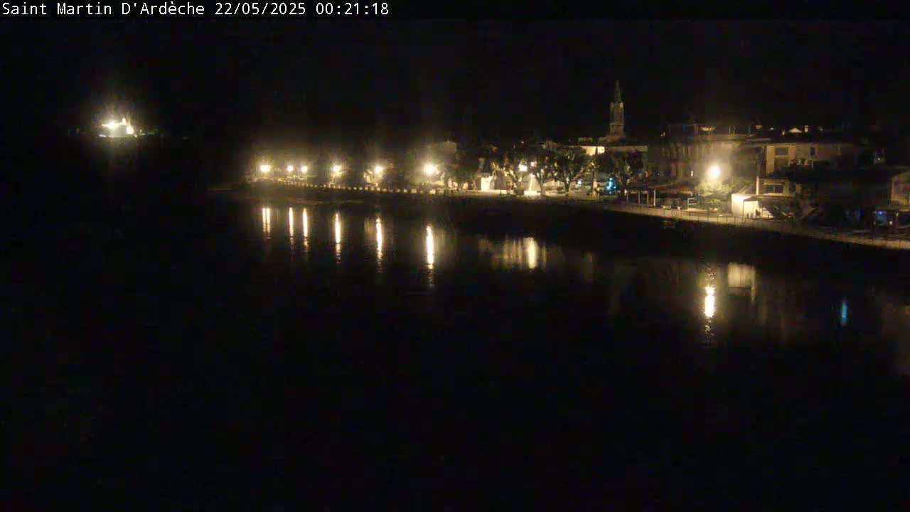 A nighttime view of a town situated alongside a calm body of water, illuminated by streetlights reflecting on the water's surface.
