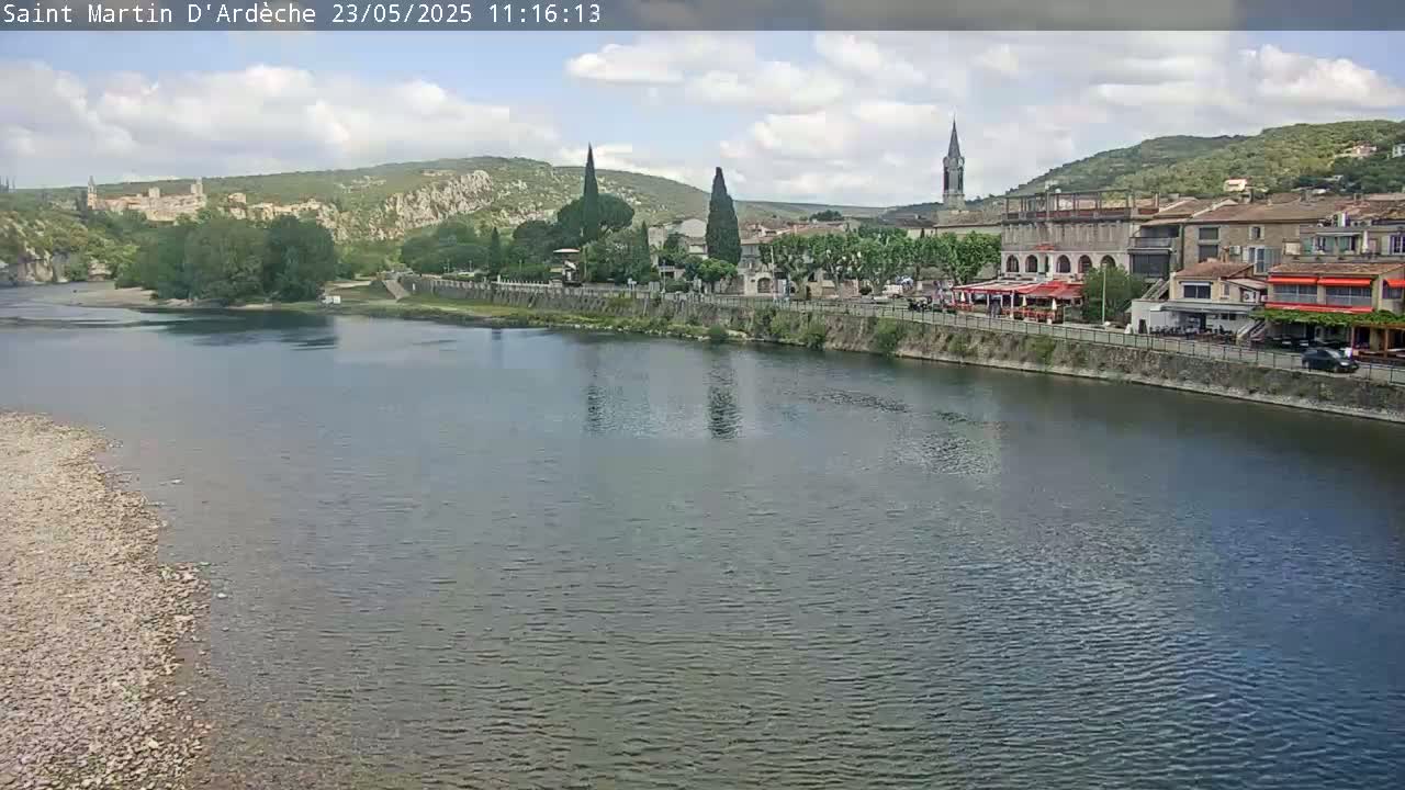 A partly cloudy day shows a river flowing past a town nestled beside hills, with buildings lining the riverbank.