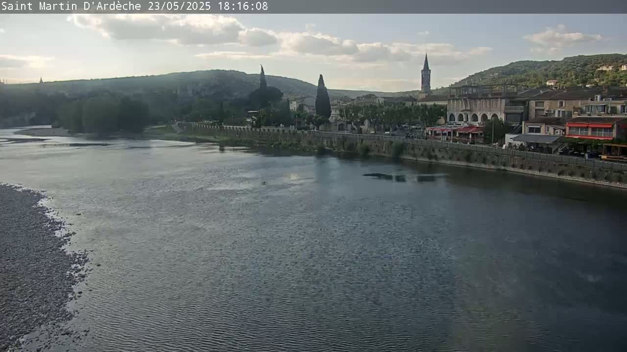 A river flows past a town nestled in a valley under a partly cloudy sky.